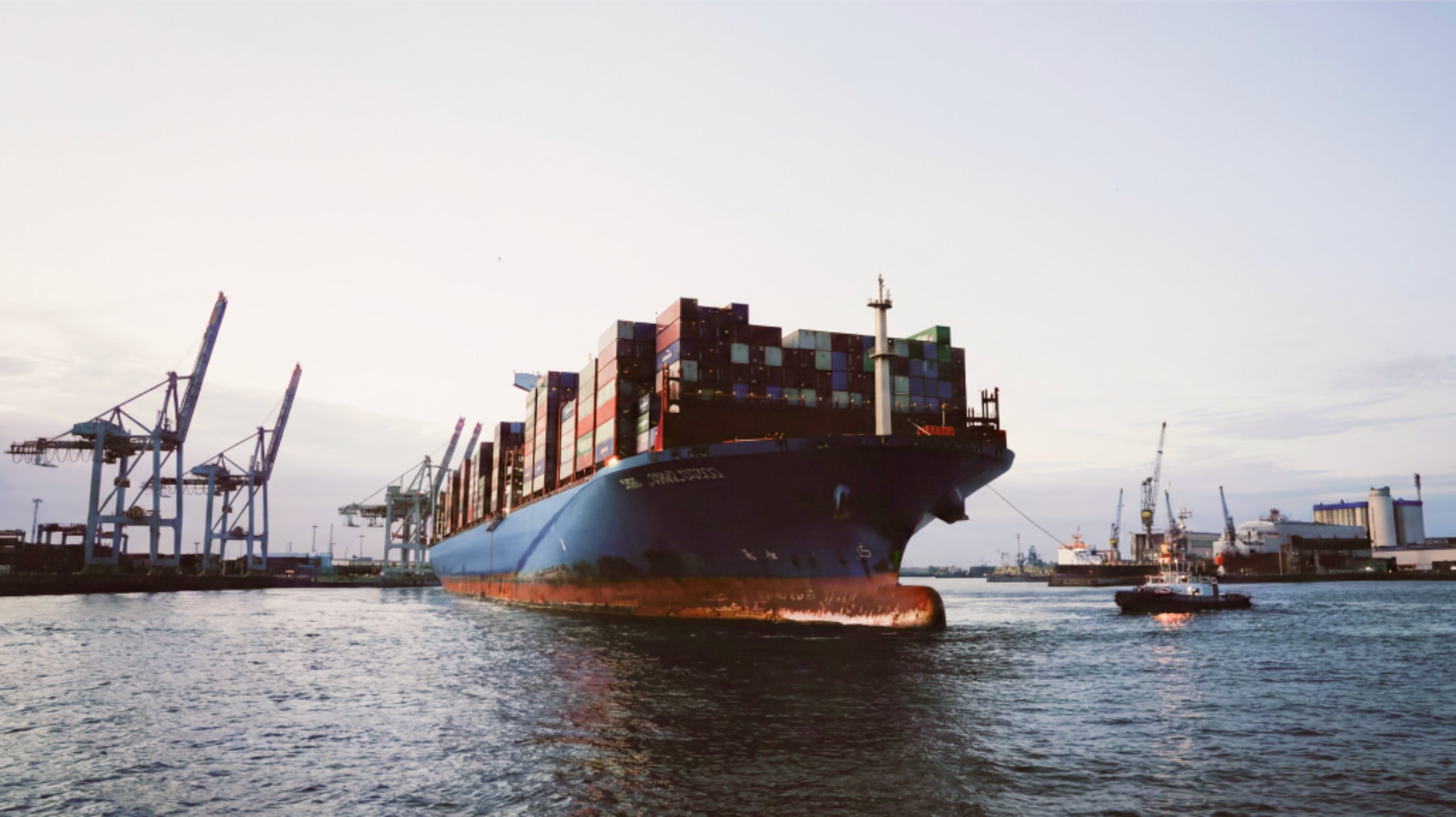 A loaded container ship leaving port at dusk, tug alongside